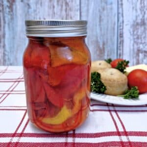 A jar of roasted red peppers on a red and white striped tablecloth, with a plate of pork pies, cheese and tomatoes in the background,.
