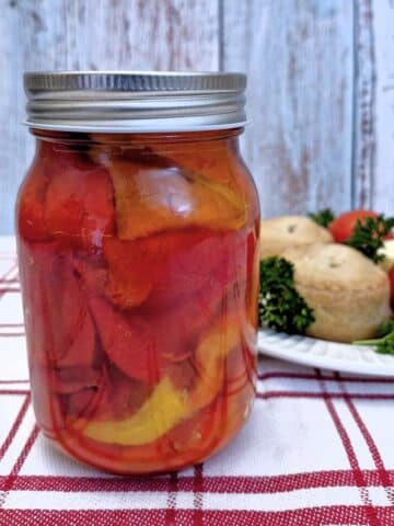 A jar of roasted red peppers on a red and white striped tablecloth, with a plate of pork pies, cheese and tomatoes in the background,.