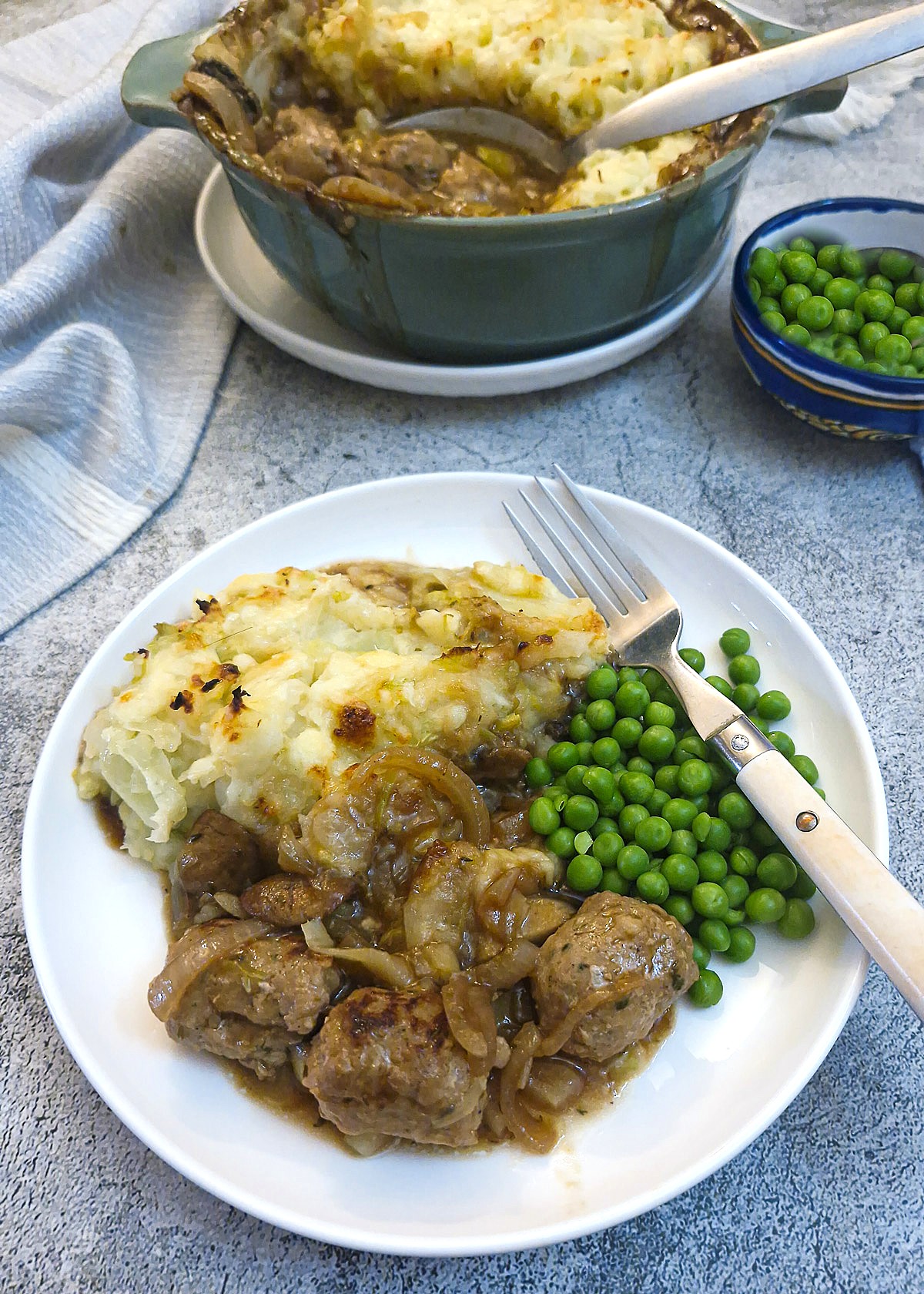 A helping of sausage and onion casserole on a white plate with mash and peas. The casserole dish is in the background.