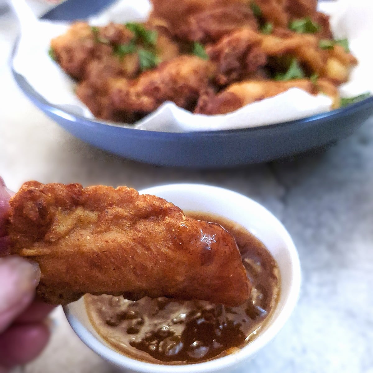 A chicken bite being dunked in a dish of dipping sauce. There is a plate of chicken bites in the background.