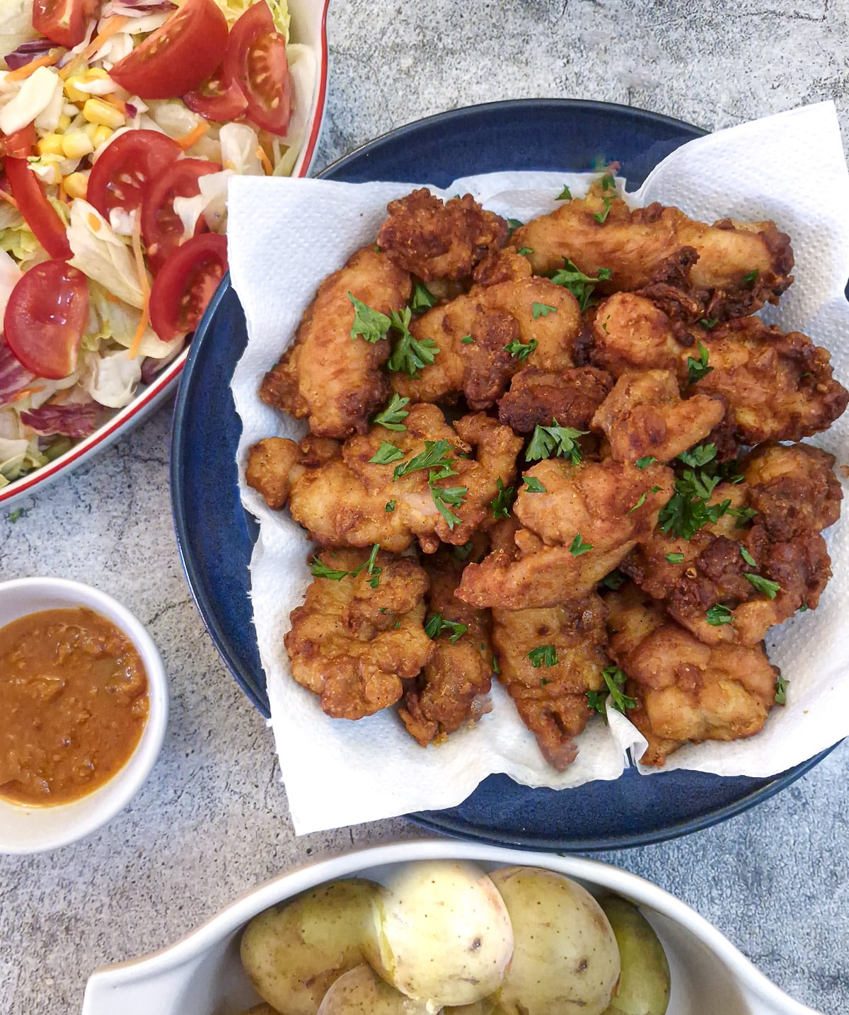A plate of chicken thigh bites, alongside a dish of salad and a bowl of boiled potatoes.