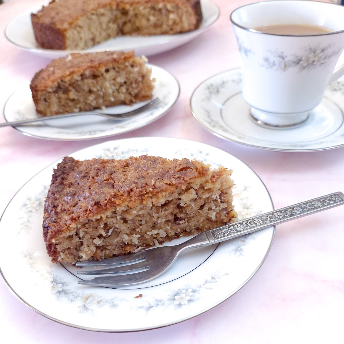 Close up of a slice of lemon and walnut coconut cake, with another plate of cake and a cup of tea in the background.
