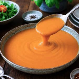 A bowl of roasted red pepper tomato cooking sauce. A largee spoon is being used to lift some of the sauce from the dish.