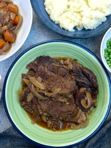A green serving dish filled with slices of slow cooked beef. There are dishes of vegetables surrounding it.