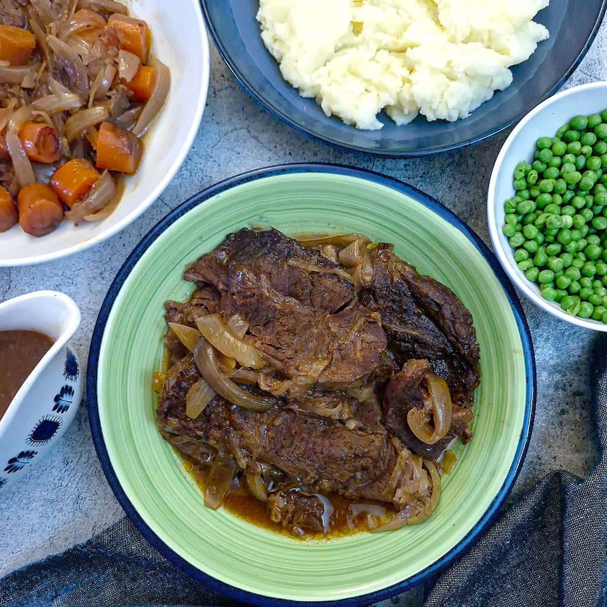 A green serving dish filled with slices of slow cooked beef. There are dishes of vegetables surrounding it.