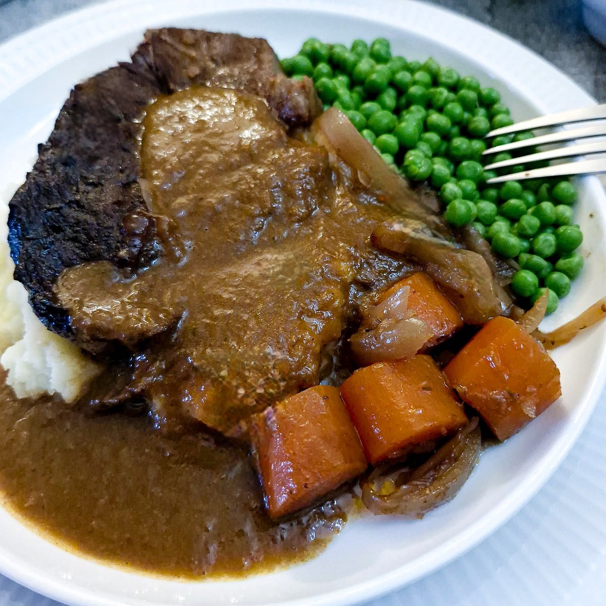 Closeup of a large piece of slow braised stewing steak on a pile of mashed potatoes, covered with gravy. There are peas and carrots on the plate.