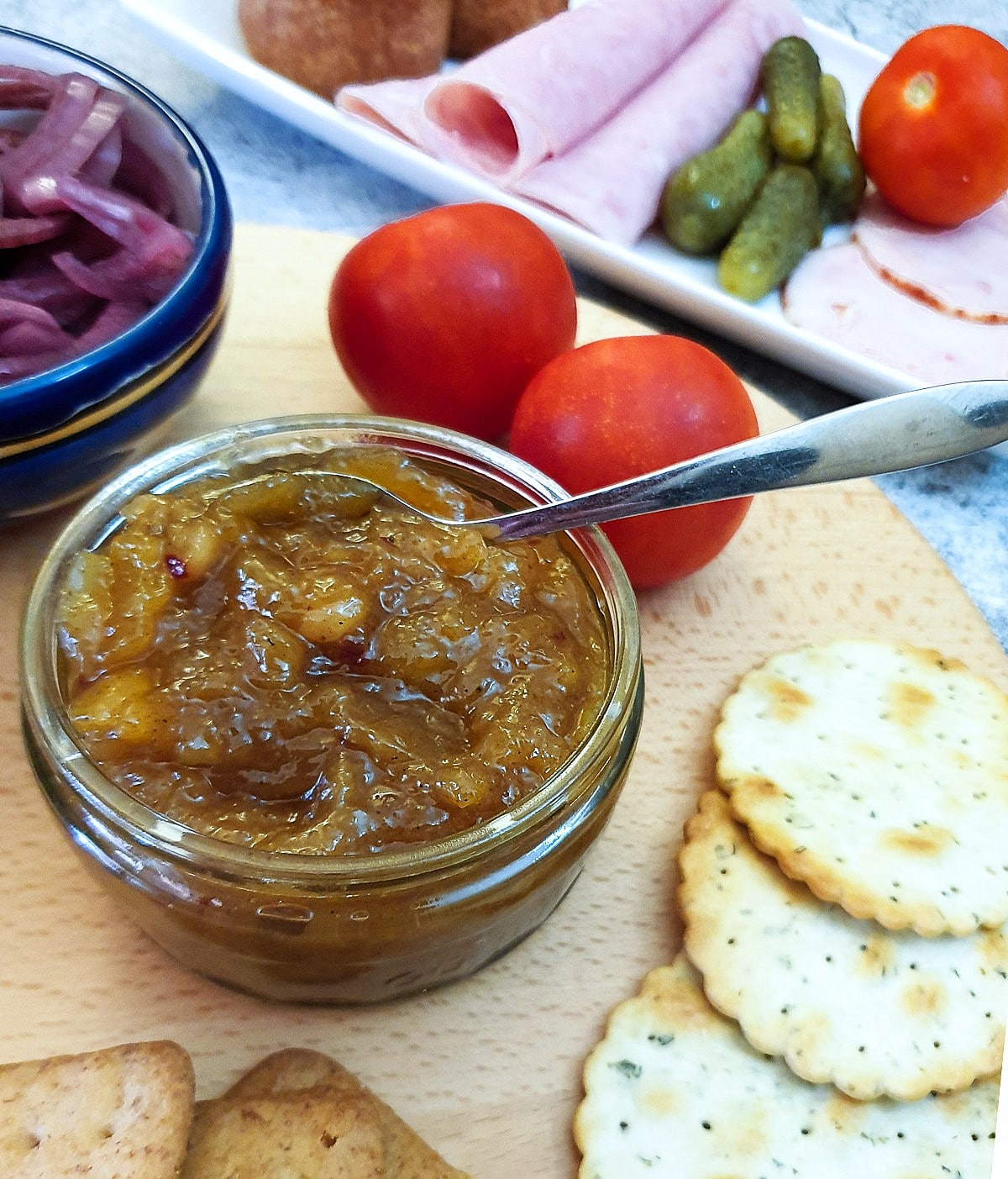 A jar of homemade chutney on a wooden board with tomatoes, ham and crackers.