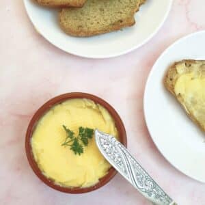 A dish of homemade butter with a sprig of parsley on top. There is a butter knfe and a plate of bread and butter alongside.