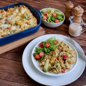 A serving of chicken and vegetable pasta bake in a white dish on a table with the baking dish in the backgrount. There is a small dish of crisp green salad on the table with salt and pepper shakers.
