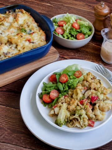 A serving of chicken and vegetable pasta bake in a white dish on a table with the baking dish in the backgrount. There is a small dish of crisp green salad on the table with salt and pepper shakers.