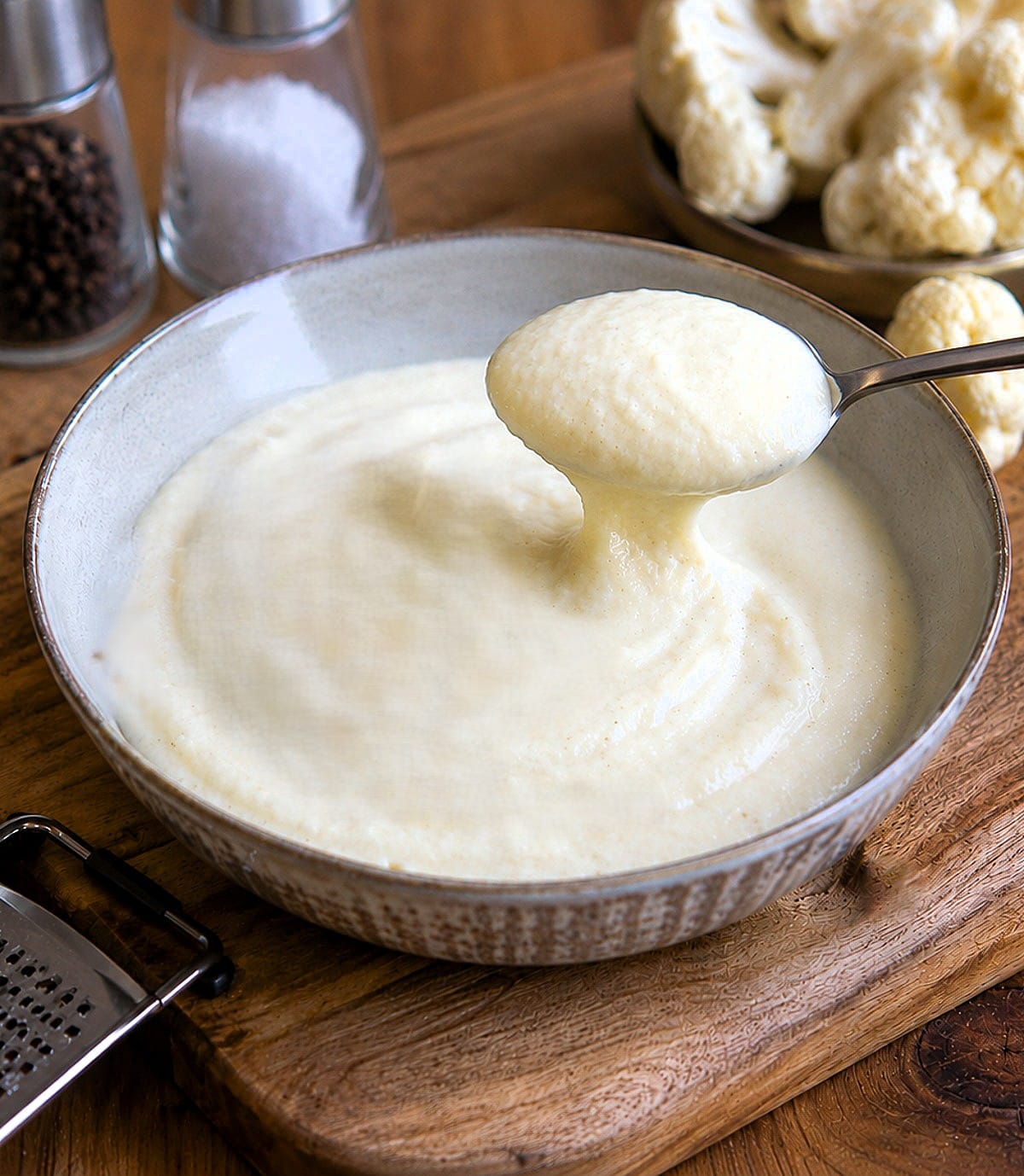 Parmesan cauliflower sauce in a grey dish on a table. A spoonful of the sauce is being lifted from the dish.