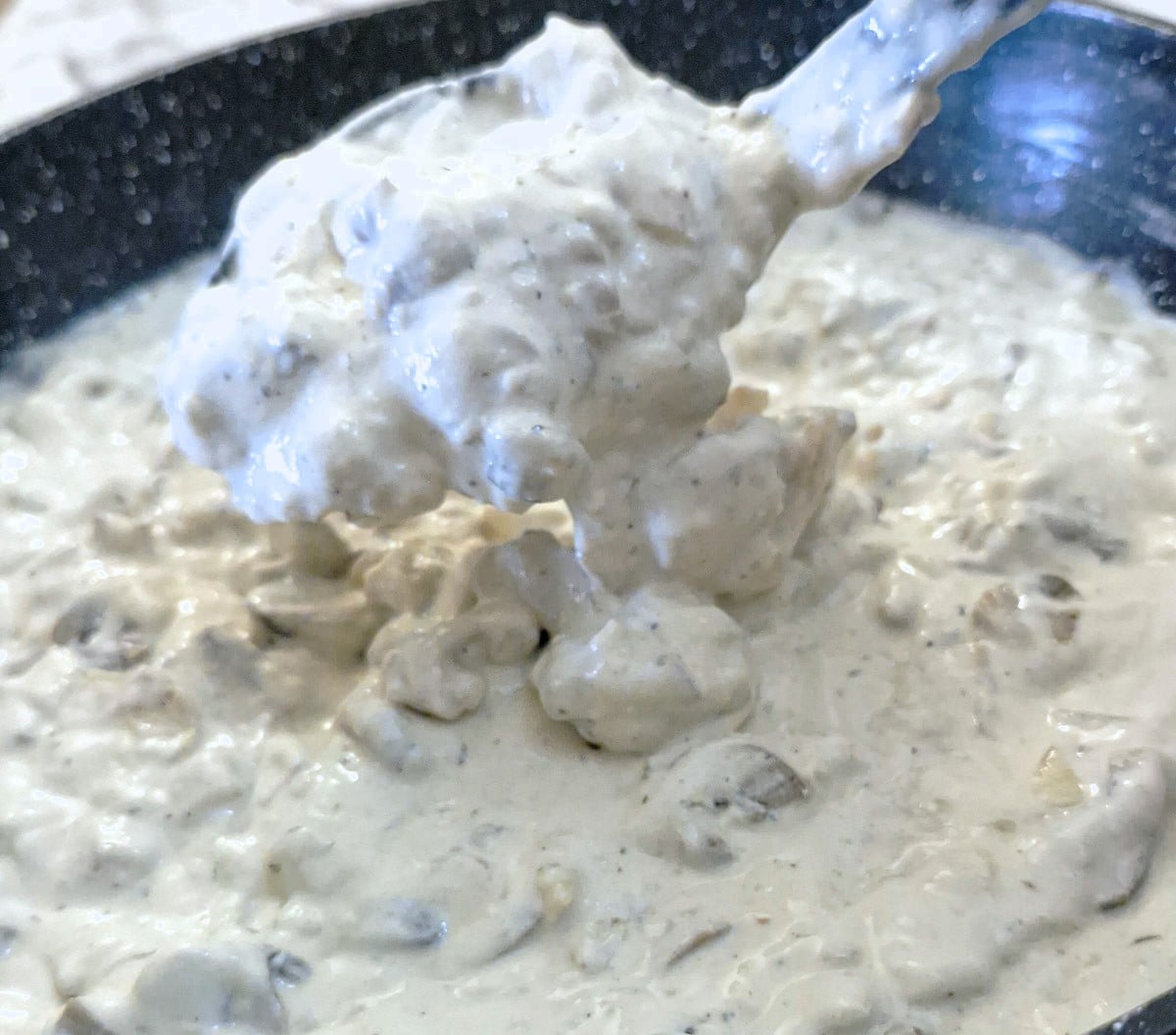 A spoonful of creamy mushroom sauce being lifted from the frying pan, showing the very thick consistency.