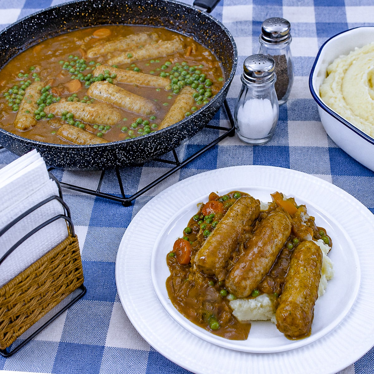 A pan of sausages in curry sauce in the background with a plate of sausages smothered in curry sauce on a pile of mashed potatoes in front.