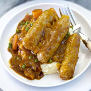 Three sausages on a pile of mashed potatoes and smothered in curried gravy. They are arranged on a white plate with a fork on the side.