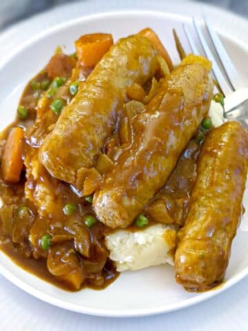 Three sausages on a pile of mashed potatoes and smothered in curried gravy. They are arranged on a white plate with a fork on the side.