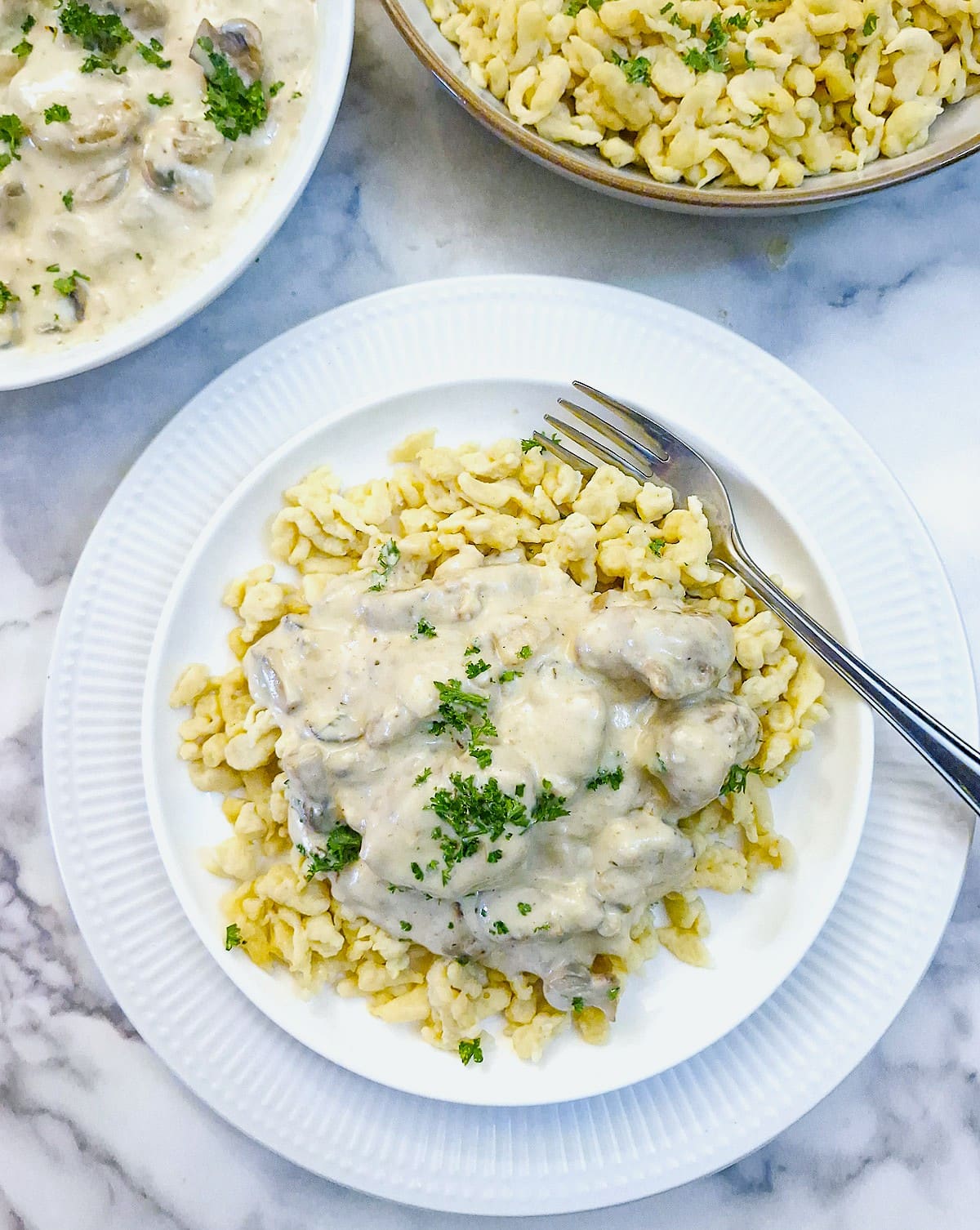 Overhead shot of rahmgeschnetzeltes on a white plate with the serving dish and a dish of spaetzle in the background.