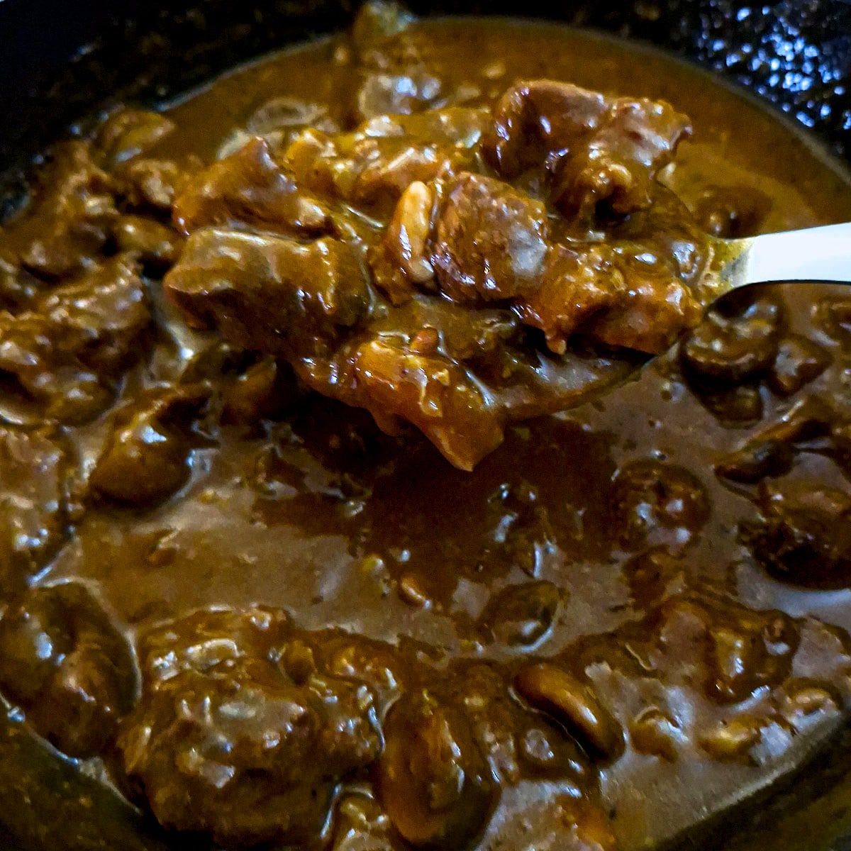 A spoonful of braised beef being lifted from the pan, dri;;ing with luscious gravy.