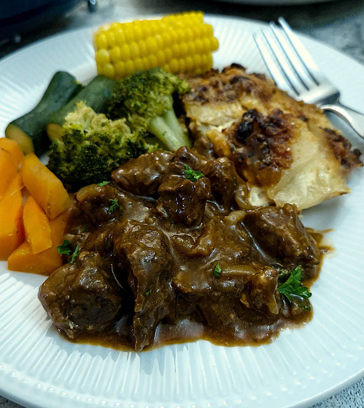 A helping of braised beef and onion on a plate with plenty of steamed vegetables.