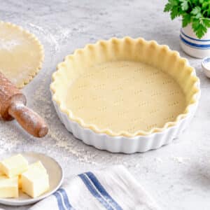 A flan dish lined with shortcrust pastry. There is a rolling pin and a dish of butter on the left hand side of the image.
