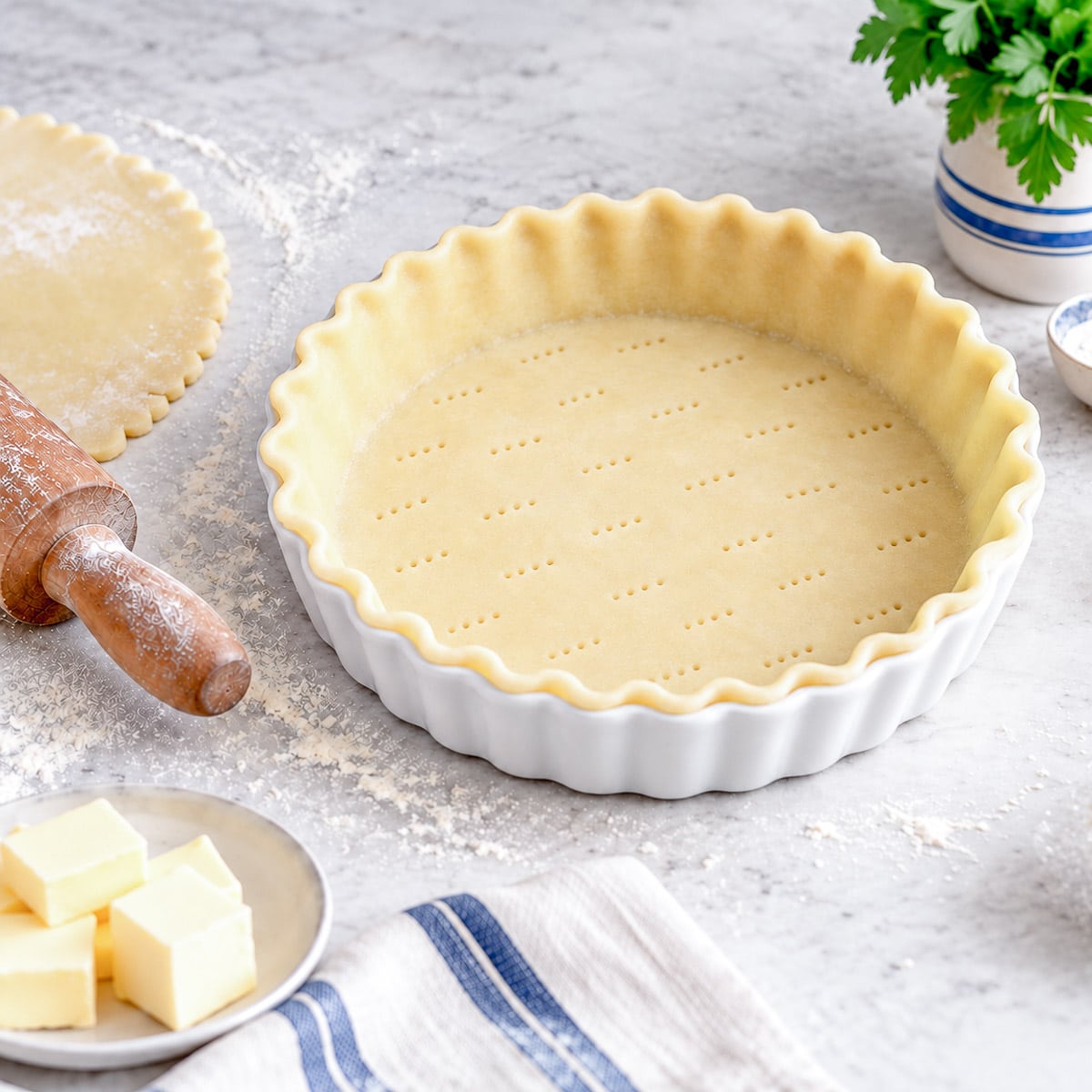 A flan dish lined with shortcrust pastry. There is a rolling pin and a dish of butter on the left hand side of the image.