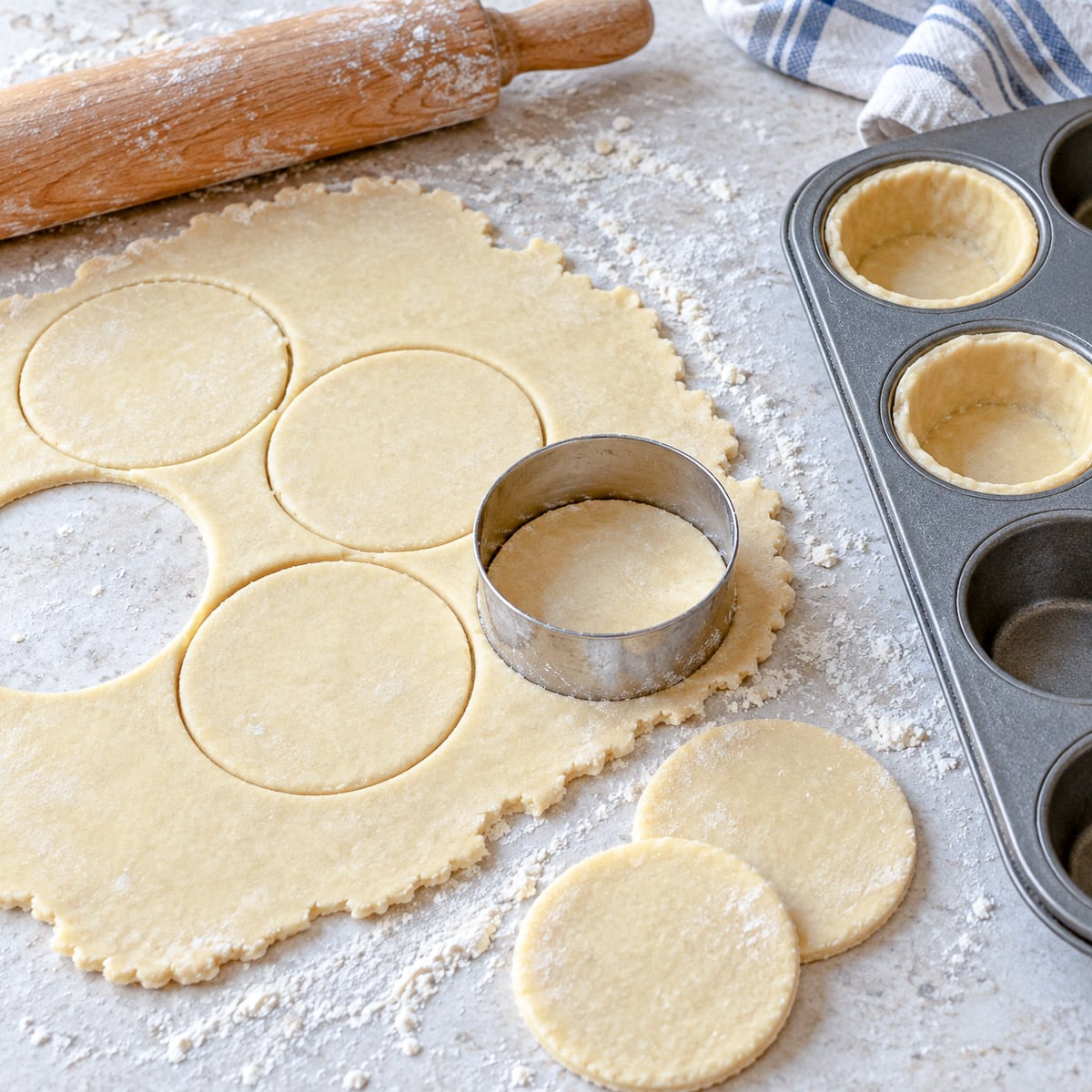 Shortcrust pastry rolled thinly on a worksurface, being cut into small rounds with a pastry cutter to line a muffin tin.