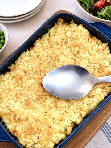 A dish of minced beef and mushroom bake topped with mashed potatoes and a layer of cheese and crushed potato crisps. There is a large serving spoon balanced on the dish.
