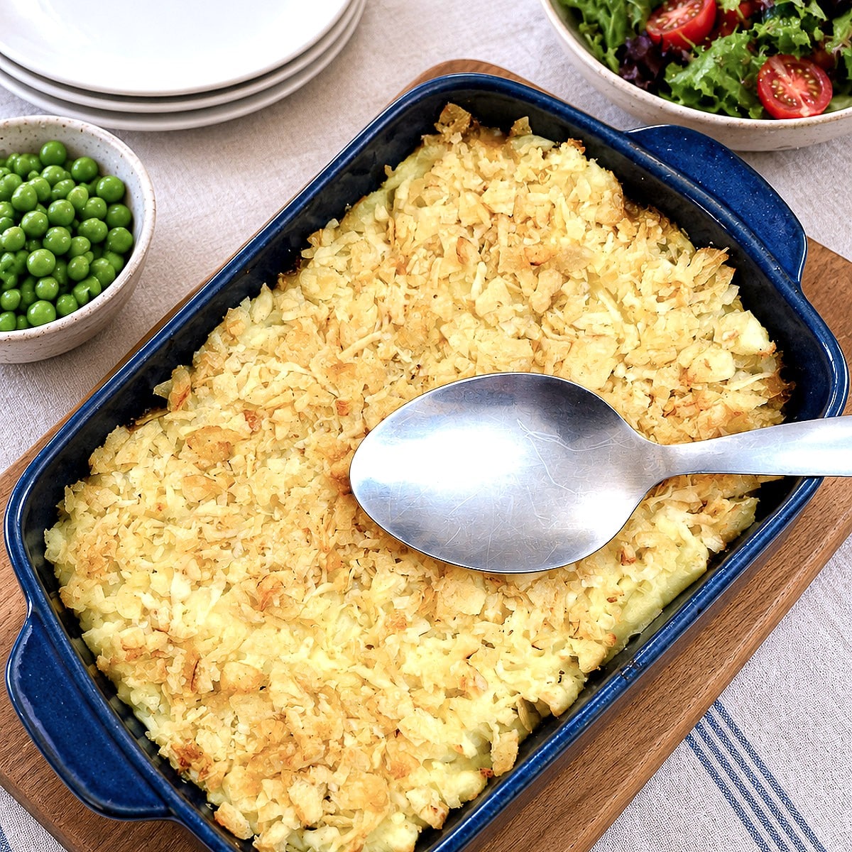 A dish of minced beef and mushroom bake topped with mashed potatoes and a layer of cheese and crushed potato crisps. There is a large serving spoon balanced on the dish.