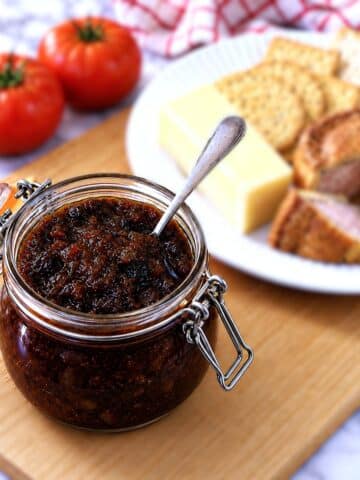 A jar of tomato chutney on a wooden bread board. There is a small spoon in the jar and a plate of cheese, crackers and pork pie in the background.