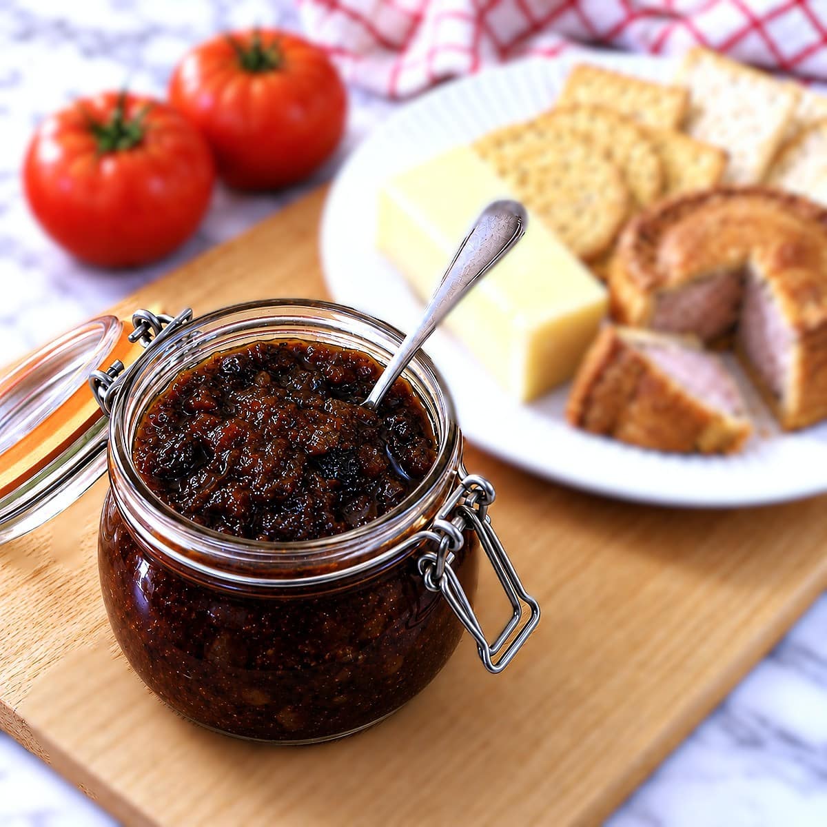 A jar of tomato chutney on a wooden bread board. There is a small spoon in the jar and a plate of cheese, crackers and pork pie in the background.