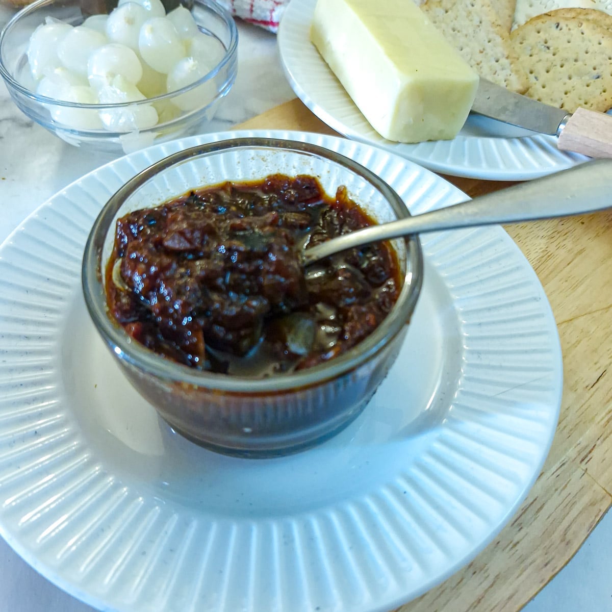 A small glass dish of tomato chutney on a white plate, with a dish of pickled onions in the background. There is a plate of cheese and crackers to one side.